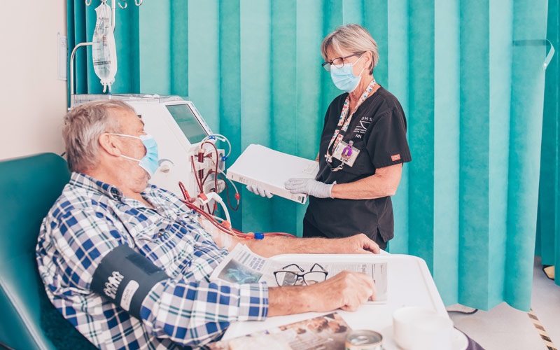 Photo of a nurse holding charts talking to older man sitting down undergoing dialysis treatment
