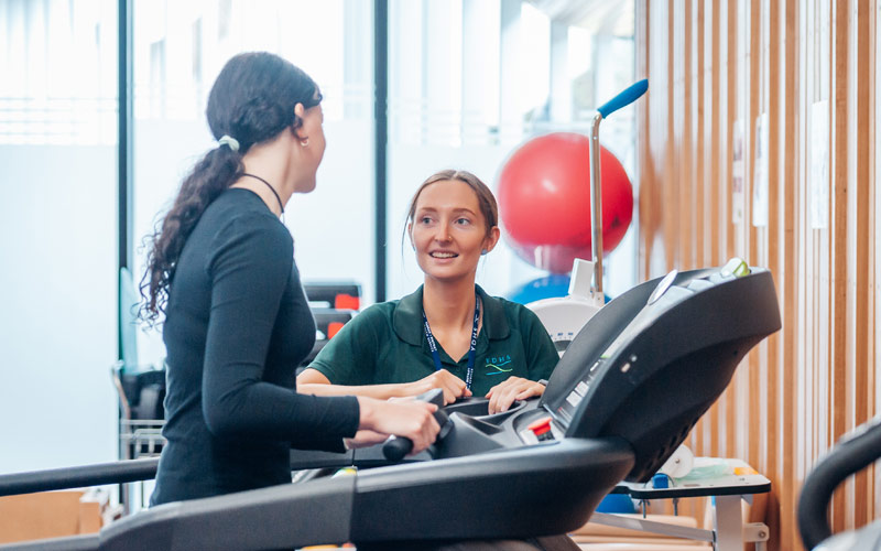 Photo of young female physio wearing dark green top, showing young female patient how to use a treadmill.