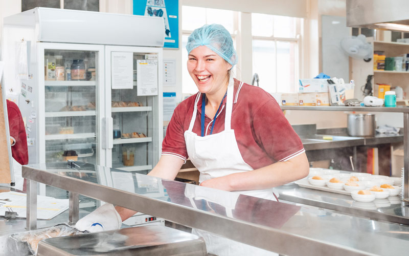 Photo of smiling girl working in nursing home kitchen wearing apron and hair net