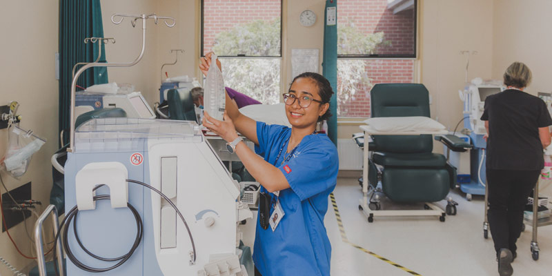 Photo of a young nurse with black hair and glasses, wearing blue uniform, changing fluids on a dialysis machine
