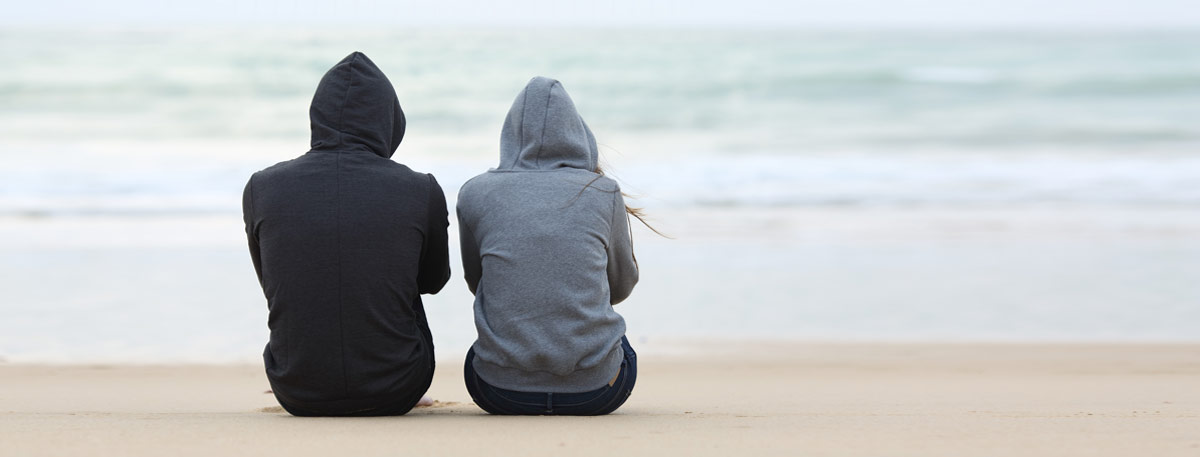 Photo of two people wearing black and grey hoodies sitting on the sand at the beach.