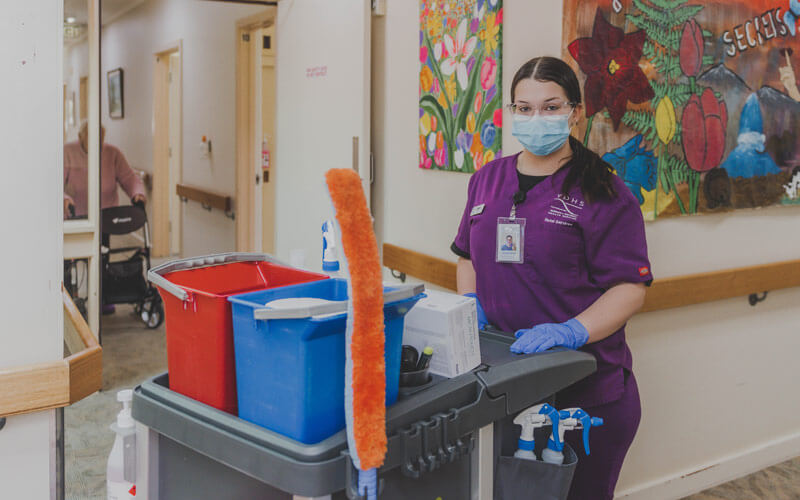 Photo of a young woman with long black hair in a pony tail, wearing a purple cleaners suit, pushing a trolley with cleaning tools and buckets. Standing in a hospital hallway.