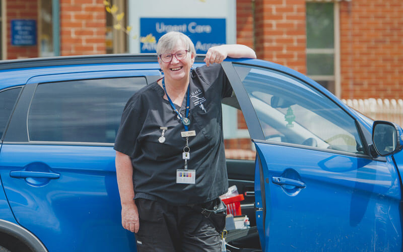 Photo of a nurse with short grey hair and wearing a black uniform, leaning against a blue car.