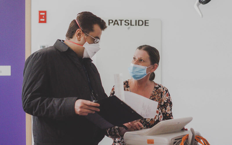 Photo of a tall male doctor wearing black and looking at a female nurse. The are both wearing masks and looking at a clipboard and paperwork.