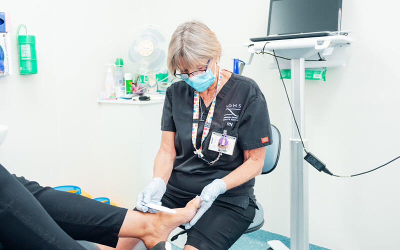 Photo of a nurse wearing black, sitting on a stool and treating the let foot of a patient.