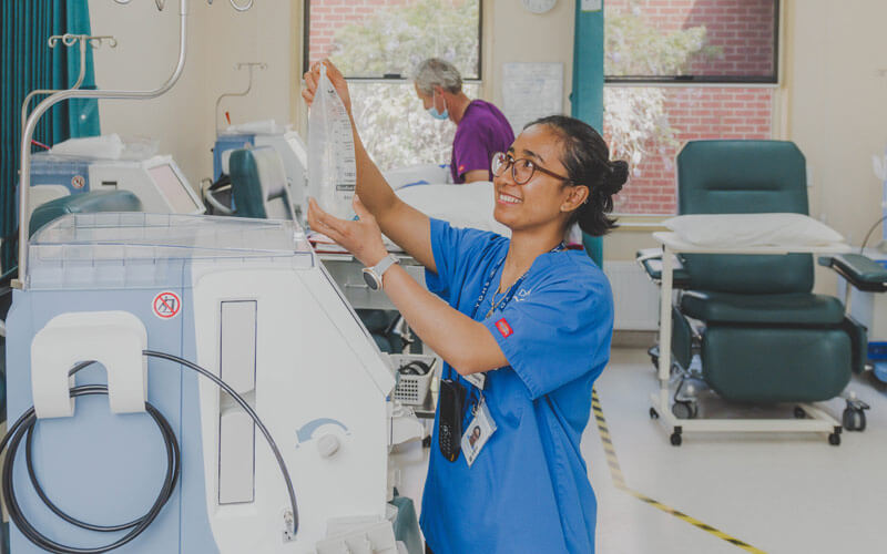 Photo of a young female nurse wearing a blue uniform, attending to a dialysis machine.