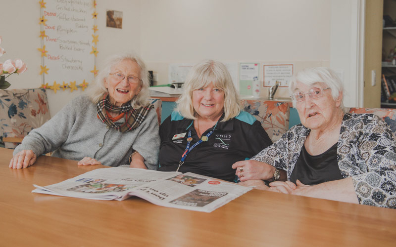 Photo of three elderly ladies sitting at a table reading the newspaper.
