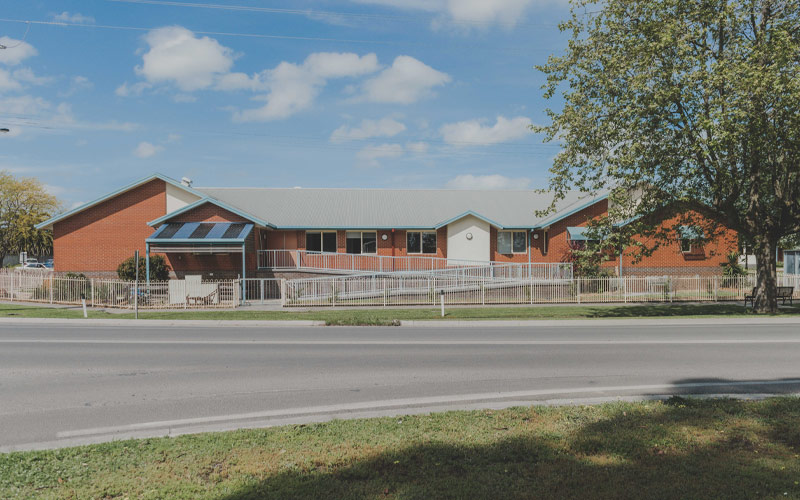 Photo of St Elmos Nursing Home from across a road, the building has red bricks, silver tin roof and lots of windows. There is a cream colour metal fence around the building.
